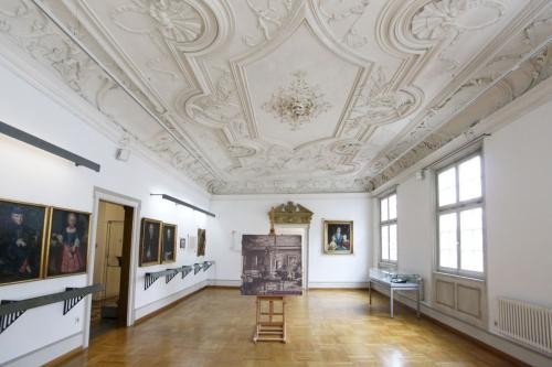 großer Raum mit Stuckdecke, links Bilder an der Wand und eine Tür, rechts Fenster und Heizkörper, in der Mitte des Raumes steht eine Staffelei mit einem historischen Bild