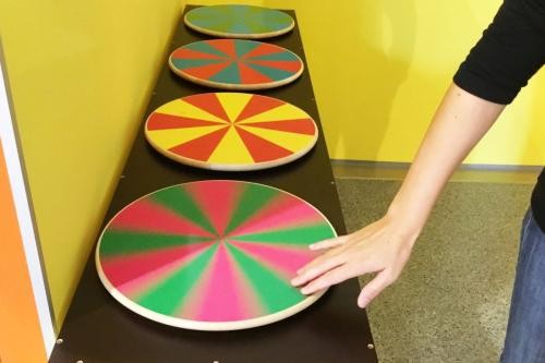 Tisch mit vier runden Platten, auf denen jeweils zwei Farben abwechselnd angeordnet sind. Eine Hand bringt eine der Platten zum Drehen.