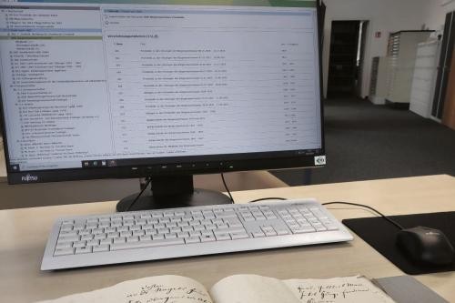 historisches Buch liegt aufgeschlagen vor einem PC. Auf dem Monitor ist ein digitales Aktenverzeichnis zu erkennen