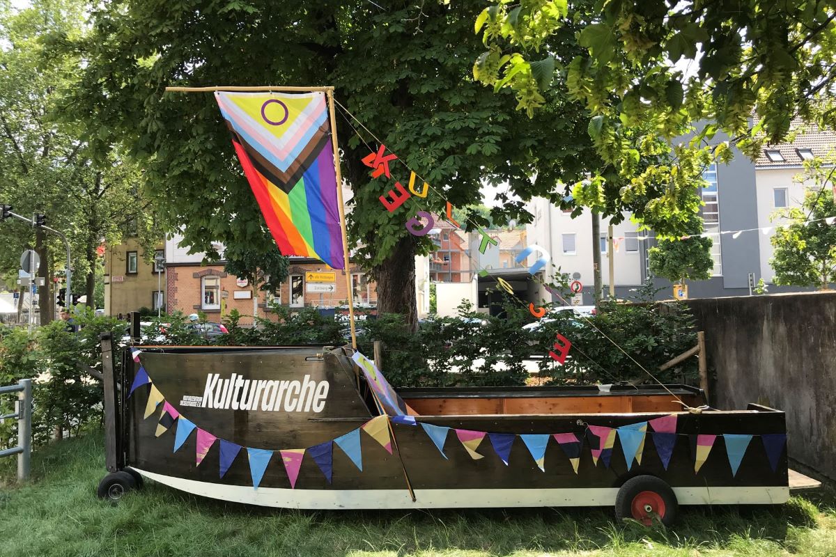 Das schwarze Holzboot "Kulturarche" steht auf einer Wiese, geschmückt mit Farbigen Flaggen und LGBTQ+ Flagge