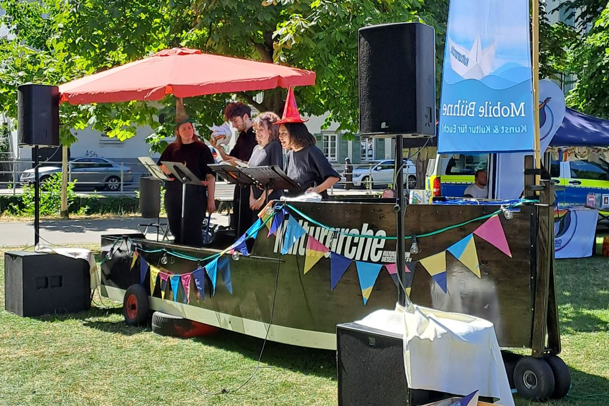 Die Kulturarche steht in einem grünen Park. Auf der mobilen Bühne, einem Holzboot, performen vier Personen an Mikrofonen unter einem roten Sonnenschirm.