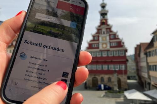 Finger zeigt auf Chatbot auf Startseite am Smartphone mit Altem Rathaus im Hintergrund