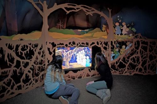 zwei Mädchen schauen auf einen Bildschirm, auf der die Geschichte der Wurzelkinder erzählt wird. Der Raum in dem sie sitzen ist wie eine Wurzelhöhle gestaltet