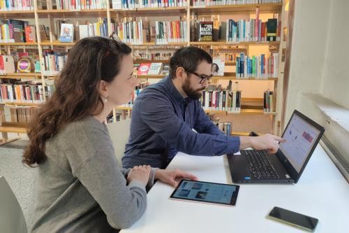 Mann mit Brille und Bart und Frau mit langen brünetten Haaren sitzen vor einem Laptop, einem Tablet und einem Smartphone. Im Hintergrund Bücherregale
