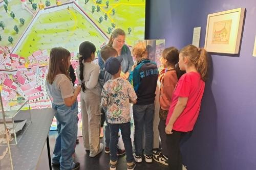 Pädagogin zeigt sieben Kindern ein Ausstellungsstück im Museum. Im Hintergrund ein großes Gemälde, rechts an der Wand ein Bild
