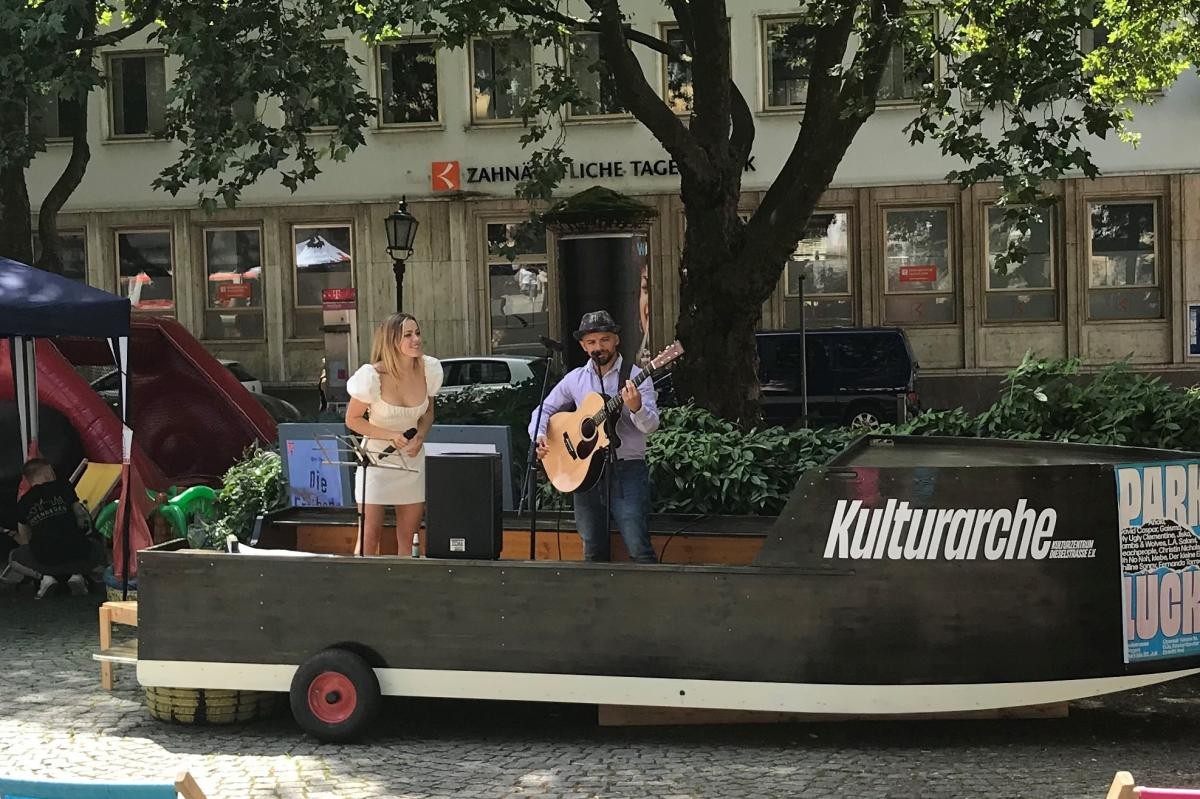Kulturarche beim Sicherheitsfestival »Auf deiner Seite« 2022 Auf dem schwarzen Holzboot "Kulturarche" steht eine Sängerin am Mikrofon neben einem Gitarristen.