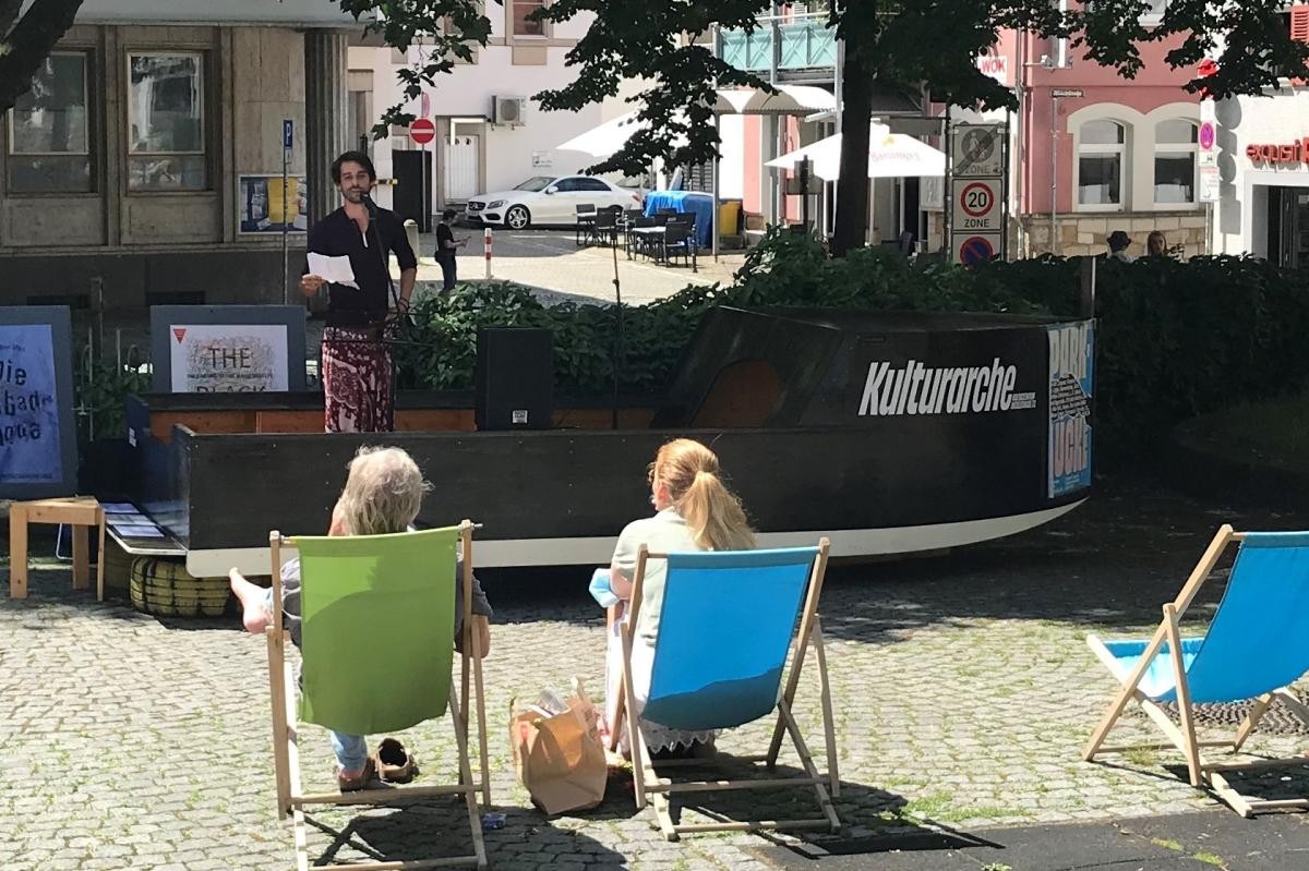 Auf dem schwarzen Holzboot "Kulturarche" steht ein Sprechkünstler am Mikrofon. Davor sitzen zwei Damen in Liegestühlen.