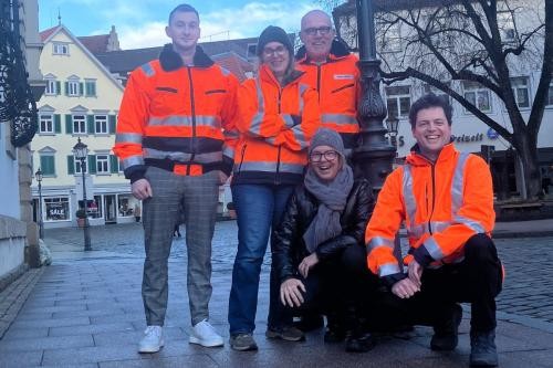 Fünf Mitarbeitende der Stadtverwaltung stehen in orangen Arbeitsjacken gekleidet vor einer Straßenlaterne in der Ritterstraße