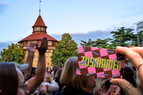 Publikum klatscht, singt und tanzt unter freiem Himmel. Im Hintergrund der Dicke Turm der Esslinger Burg. Im Vordergrund eine Hand, die eine Karte im Scheckkartenformat hält, auf der "Zusammen" steht