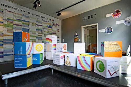 Blick in einen Ausstellungsraum, in dem bunte Würfel auf einem Podest stehen. An der Wand ist ein großer Kalender angebracht, auf der anderen Wand steht "Gebet"