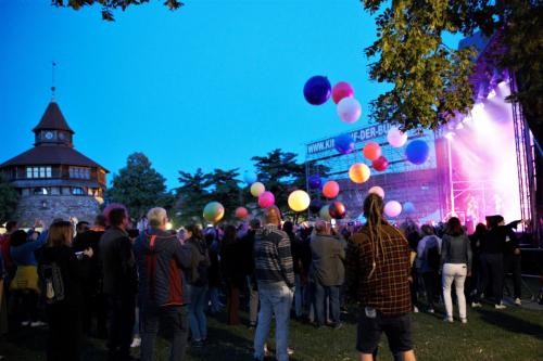 Menschen auf einer Wiese im Freien. Links der Dicke Turm der Esslinger Burg. Rechts eine Konzertbühne mit Scheinwerferlicht. Der Himmel ist nachtblau. Über dem Publikum schweben bunte Bälle.