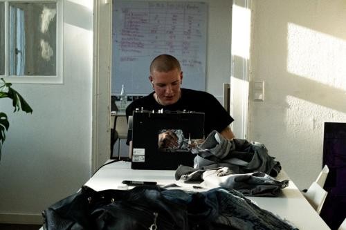 Junger Mann sitzt an Stirnseite eines langen Tisches an einer Nähmaschine