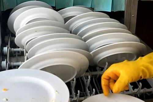 Hand in gelbem Gummihandschuh räumt Teller in eine Industriespülmaschine ein