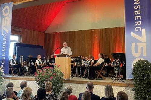 auf einer Bühne in einer Halle steht ein Mann am Redepult vor Publikum. Hinter dem Redner sitzt ein Orchester
