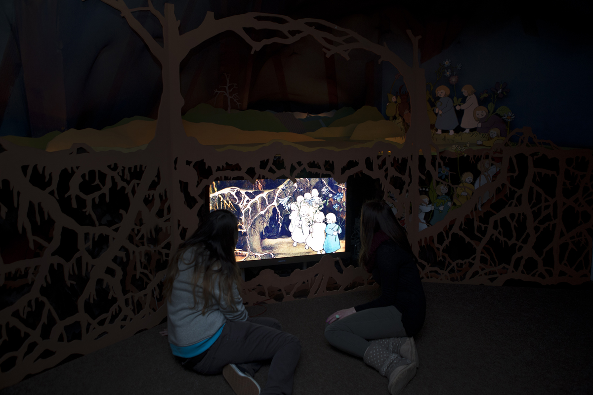 In der dunklen Wurzelhöhle sitzen zwei Besucherinnen vor einem erleuchteten Bild der Wurzelkinder.