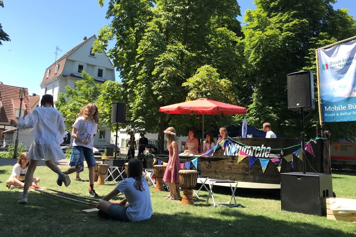 Ukrainische Gruppe "Rhythmus bewegt" von buntES auf dem Schwörfest 2023 Das Holzboot Kulturarche steht unter einem roten Sonnenschirm in einem Park. Davor tanzen junge Menschen