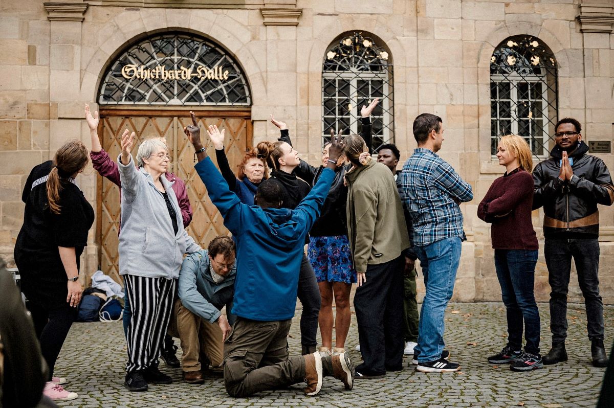 Eine Gruppe von Menschen unterschiedlichen Alters steht vor einem historischen Gebäude und führt gemeinsam eine Art Bewegungs- oder Theaterübung aus, wobei mehrere Personen die Arme nach oben strecken, während andere zuschauen oder sich abwenden.