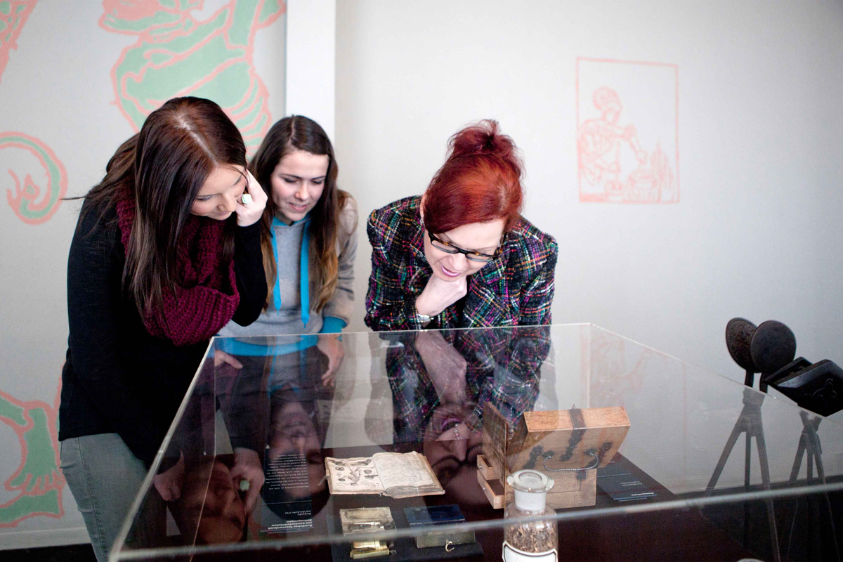 Drei Besucherinnen beugen sich über eine Vitringe und betrachten ein aufgeschlagenes Buch und ein geöffnetes Holzkästchen.