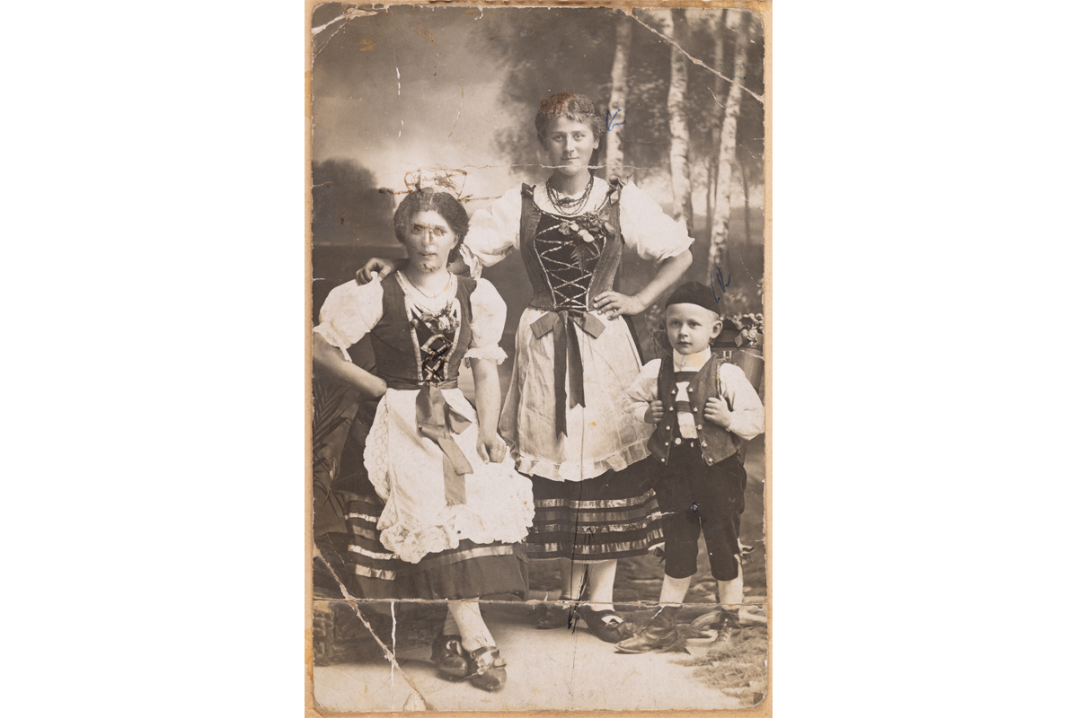 Alte, bestoßene Fotografie von zwei jungen Frauen und einem kleinen Jungen mit Bütte auf dem Rücken. Alle tragen Tracht.