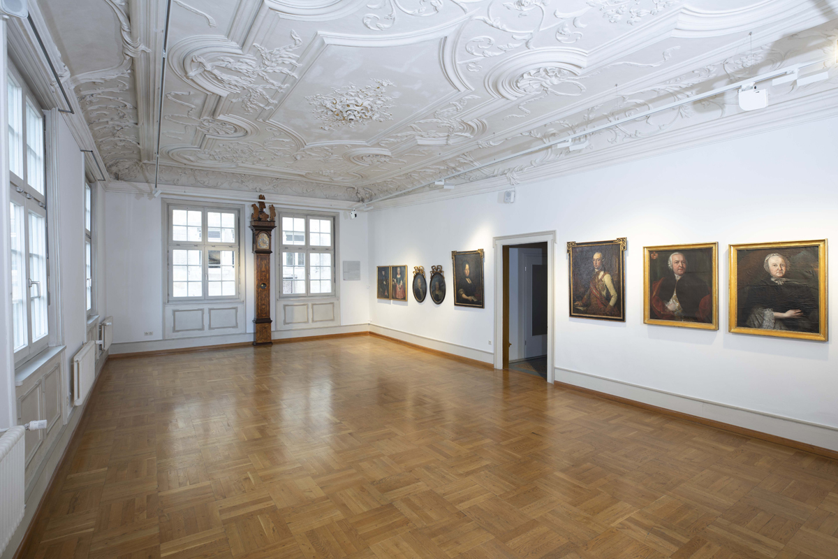 Repräsentativer Raum mit reich verzierter Stuckdecke. An den Wänden hängen Gemälde.