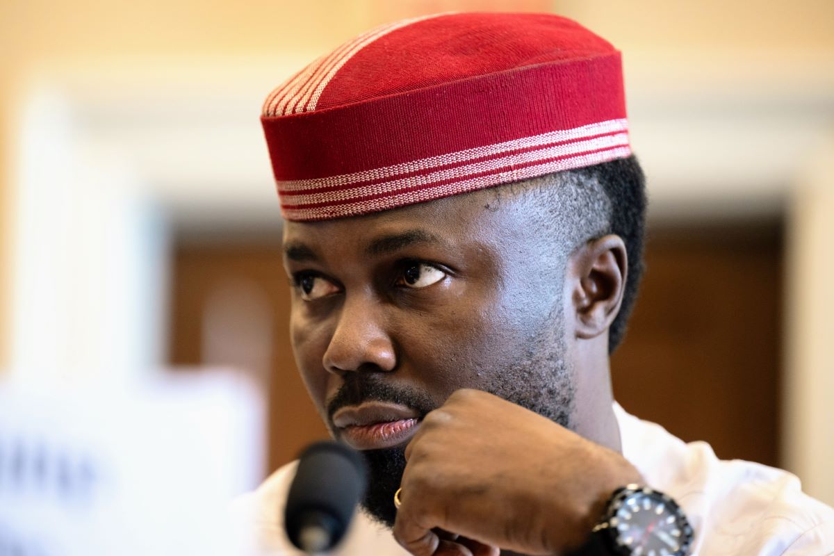 Philip Obaji jr., mit freundlicher Genehmigung der U.S. Helsinki Commission Potrait eines Mannes in weißer Kleidung und rot-weißem traditionellem Kopfschmuck, mit gepflegtem Bart, die Hand am Kinn ruhend, vor einem Mikrofon in einem Innenraum, aufmerksam und nachdenklich.