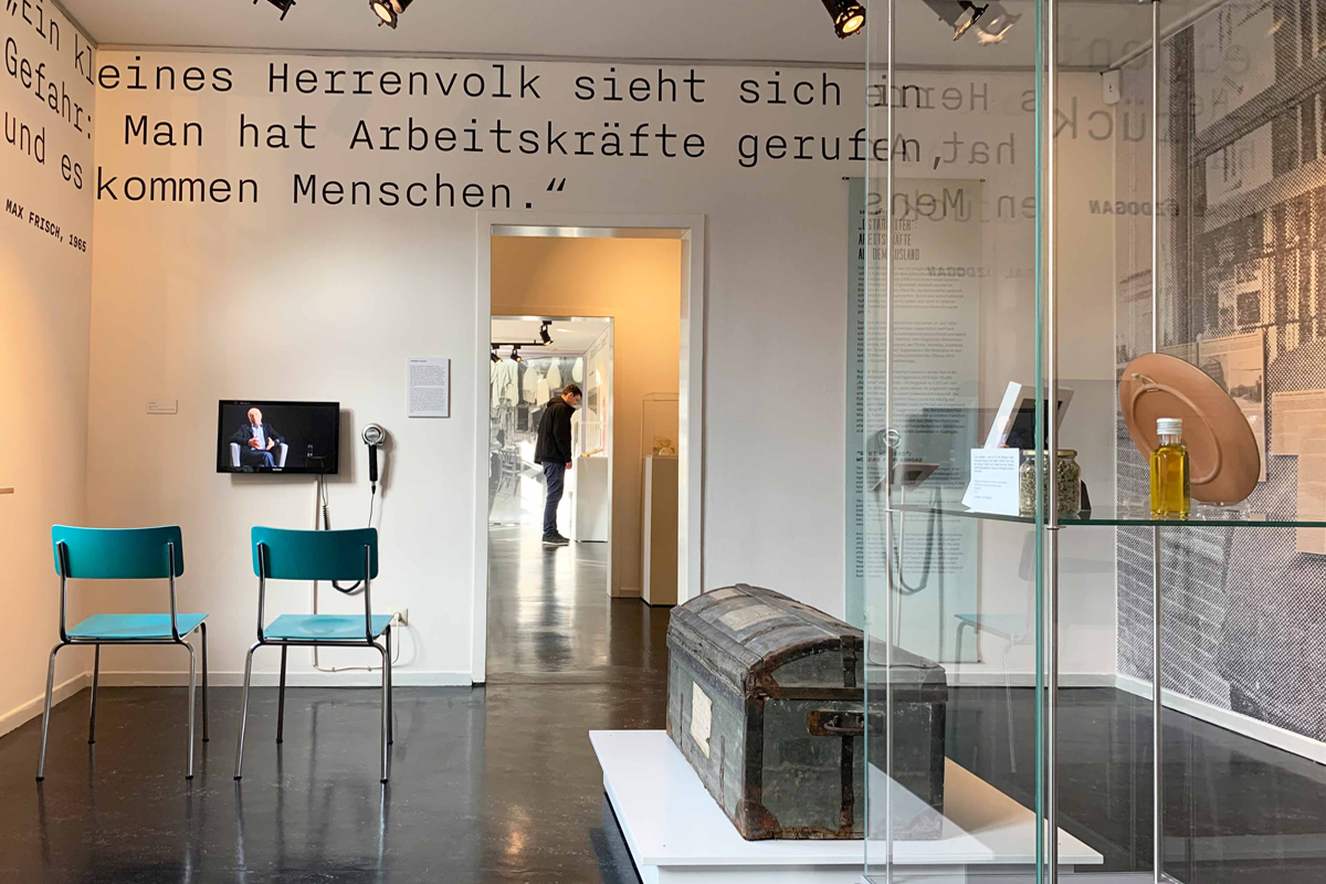 Durchblick in drei Räume. Im vordersten stehen eine Vitrine, eine alte Truhe und Stühle vor einem Bildschirm.  An der Wand sind Teile eines Zitats lesbar. "Man hat Arbeitskräfte gerufen ... kommen Menschen."