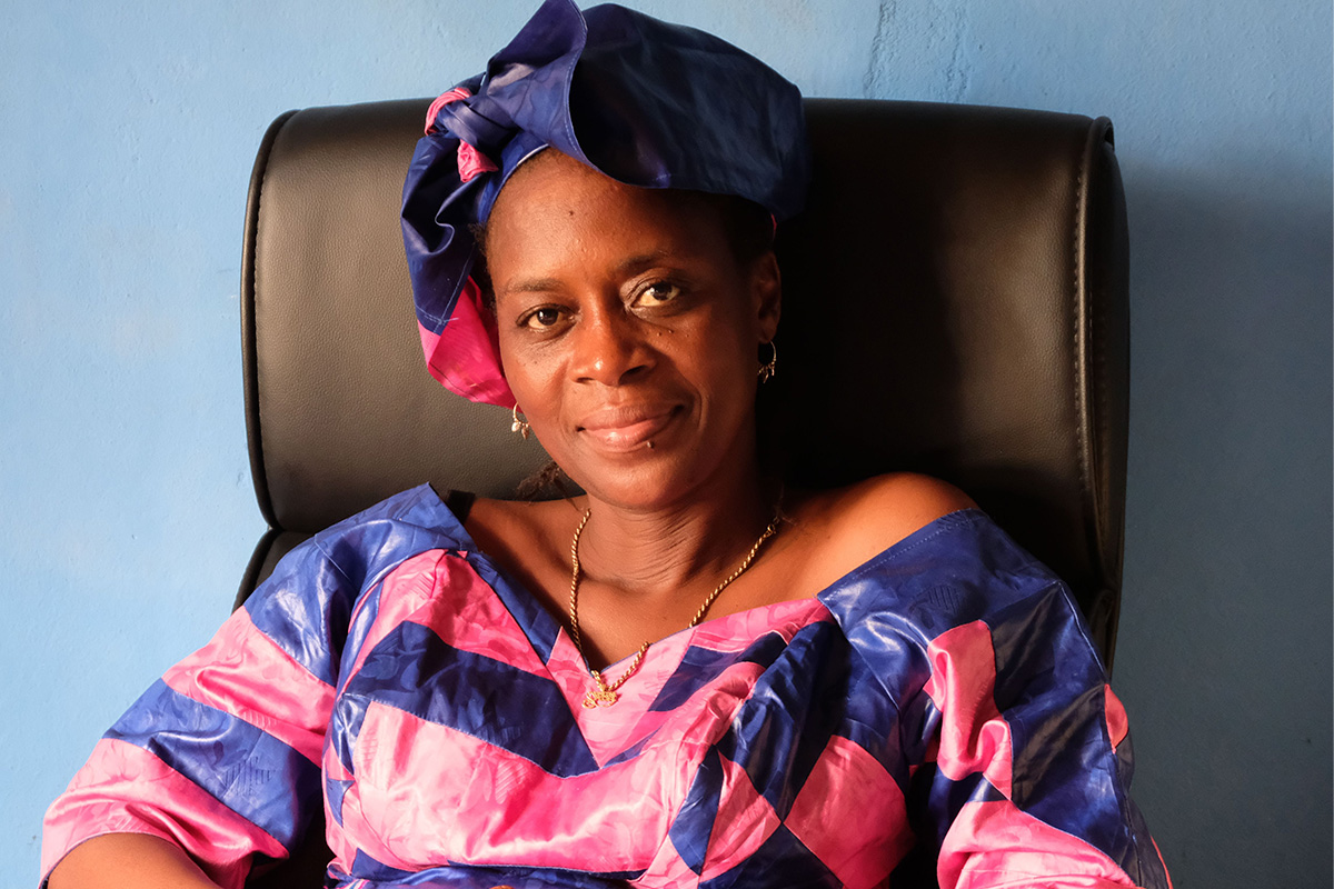 Portrait von Rugiatu "Neneh" Turay, einer Frau mit dunklem Teint in rosa-violetter Kleidung und passendem Turban, auf einem Stuhl sitzend