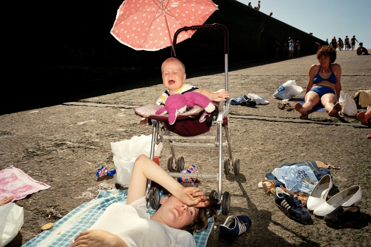 betonierter Strand, eine Frau liegt vor einem Buggy, in dem ein schreiendes Baby sitzt