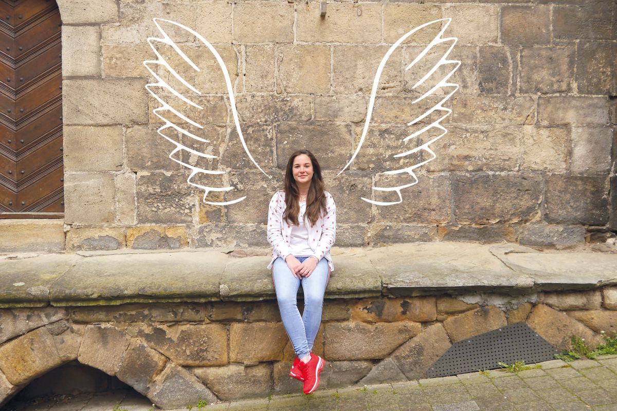 Engel Junge Frau sitzt vor einer Mauer, auf die große Engelsflügel gezeichnet sind.