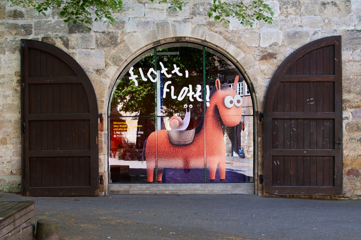 Schwörhaus von außen, das große Fenster ist beklebt mit Ausstellungstitel und einer Illustration: ein Pferd, auf dem eine Schnecke sitzt.