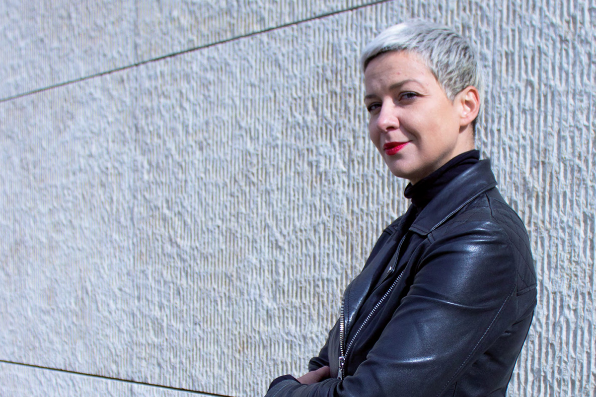 Portrait von Maria Kalesnikava: Eine junge Frau mit graublondem Pixiecut, roten Lippen und in schwarzer Lederjacke blickt vor einer hellen Mauer stehend seitlich in die Kamera