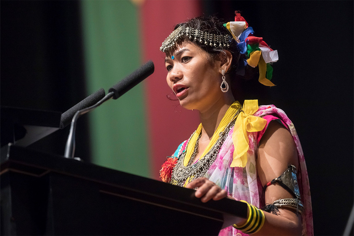Frau in farbenfroher Kleidung und auffälligem Silberschmuck auf dem Kopf, um den Hals und am Oberarm spricht am Rednerpult in ein Mikrofon