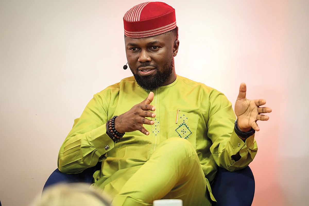 Portrait eines Mannes in hellgrüner Kleidung und mit roter Kopfbedeckung, mit einem Headset-Mikrofon und mit den Händen sachlich gestikulierend