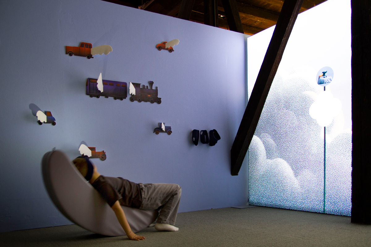 Ausstellungsraum 2: Mitmachstationen: Ein Kind liegt auf einem Polster und chillt vor einem großen Bild einer Wolke.
