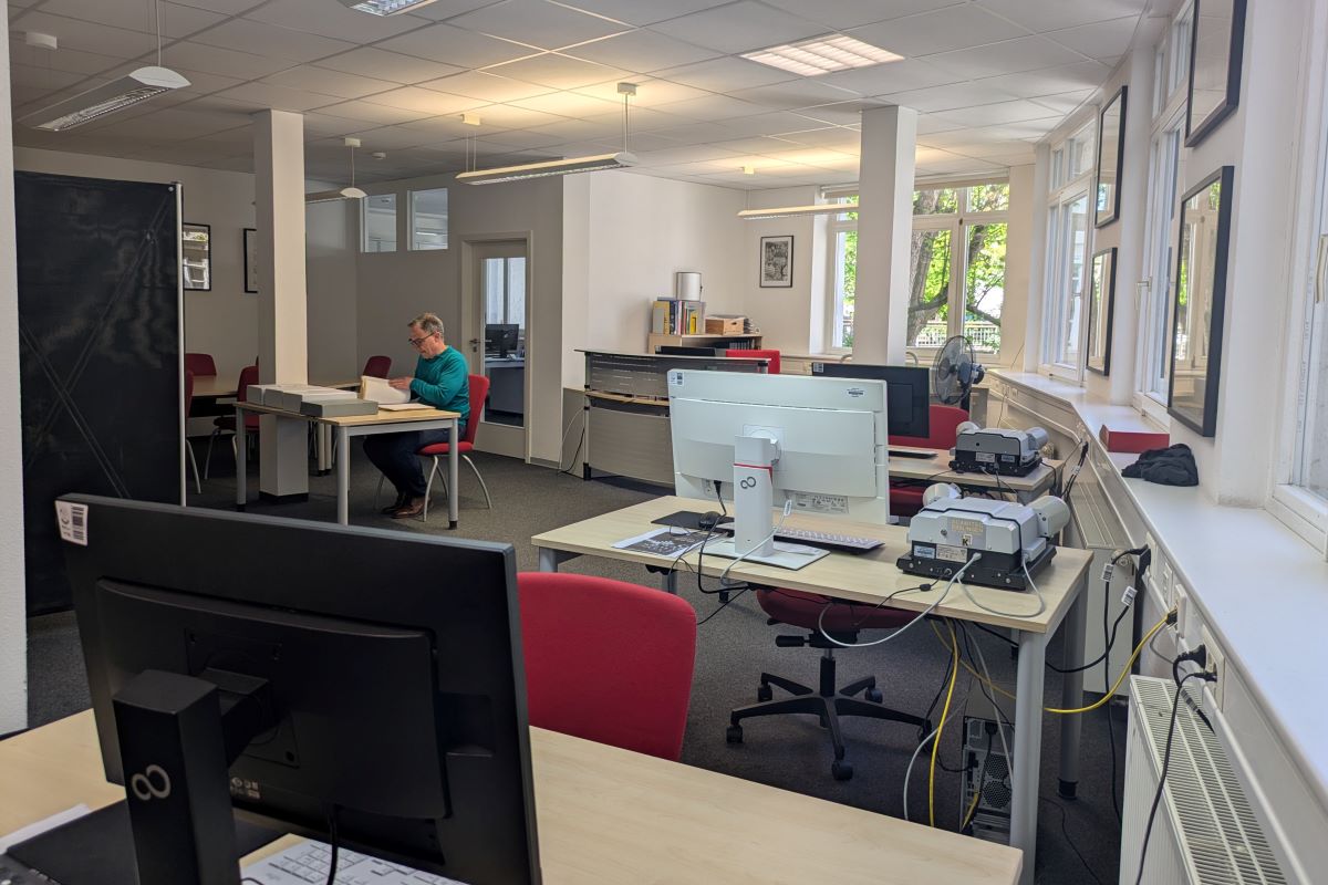 Heller Arbeitsraum mit Schreibtischen, Monitoren und Scannern; im Hintergrund sitzt eine Person und arbeitet mit großen Büchern.