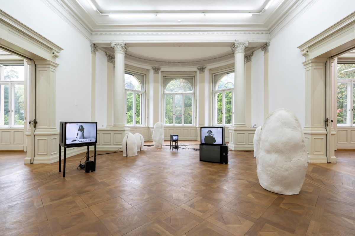 Marie Zbikowska, Mutterform, 2024 © Frank Kleinbach Blick in Saal der Villa Merkel, Parkettboden, Stuckdecke mit Oberlicht, Rundbogenfenster ins Grüne, mehrere weiße Plastiken und Fernseher stehen im Raum