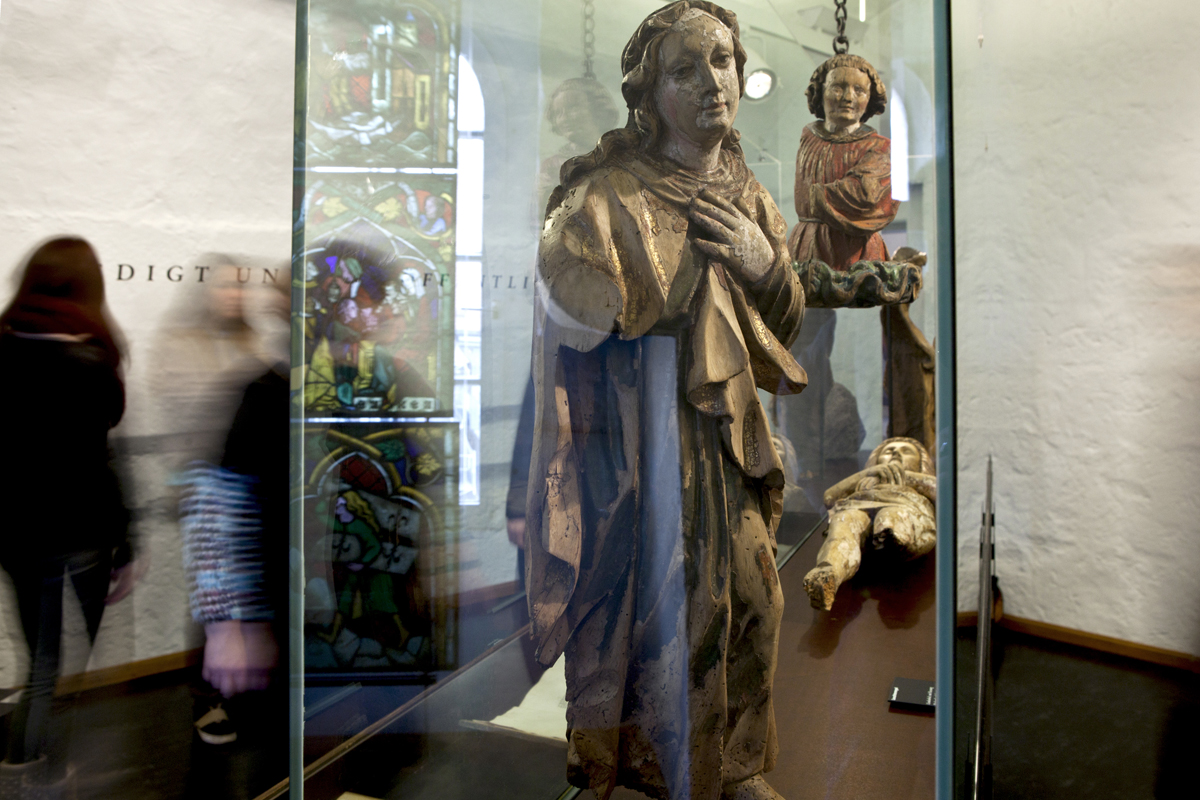 Vitrine mit religiösen Holzskulpturen: eine Heilige, ein Engel und eine liegende beschädigte Christusfigur. Im Hintergrund Besucherinnen.