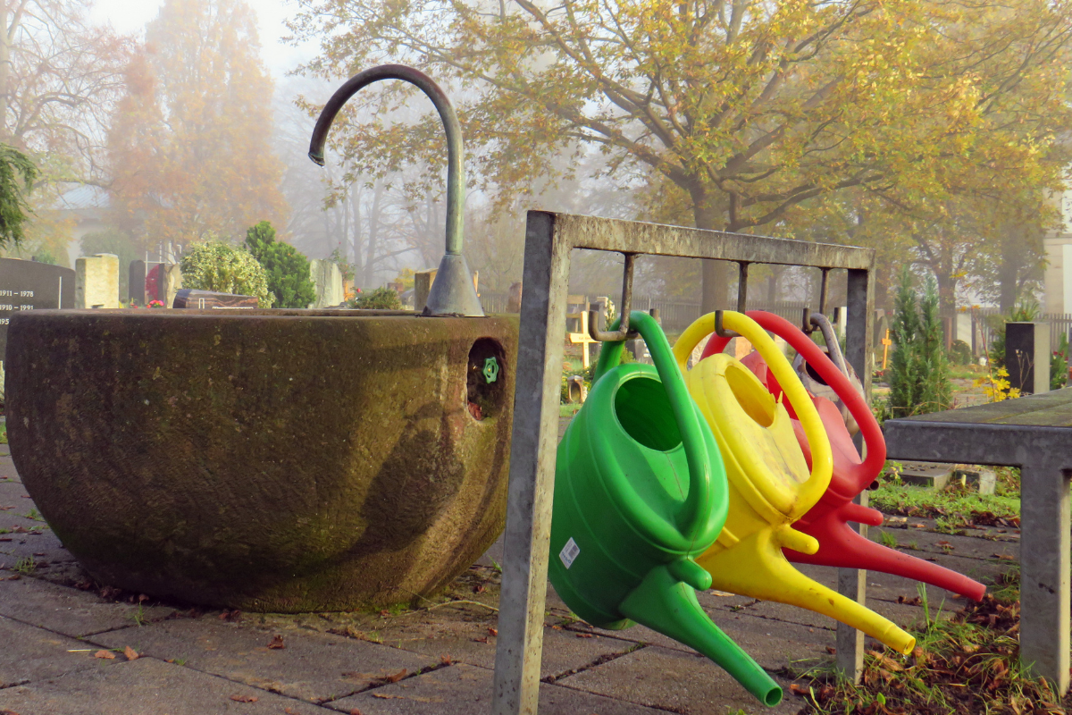 Friedhofsbrunnen Zu sehen ist ein Brunnen auf dem herbstlichen Friedhof, davor hängen Gießkannen