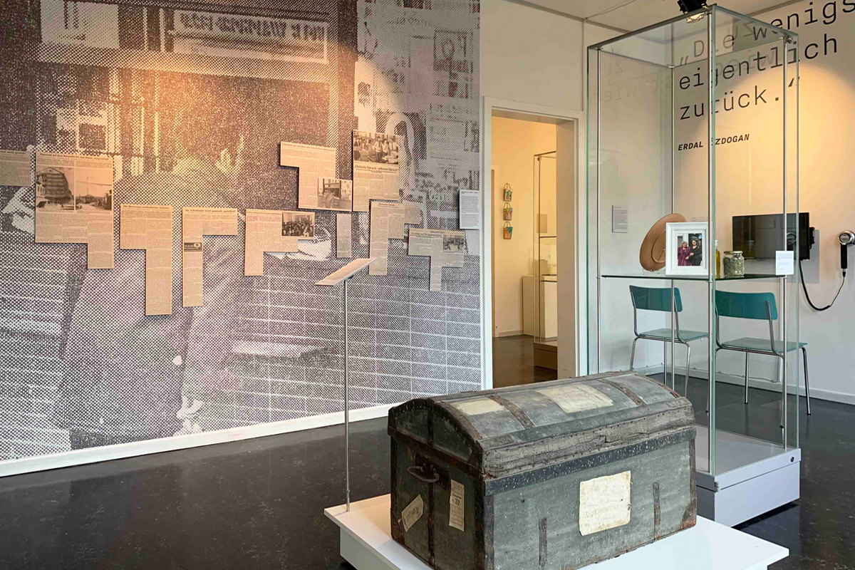 Ausstellungsraum mit einer Vitrine, Stühlen vor einem Bildschirm und einer alten Truhe. An der Wand ein großes Foto eines Mannes, der vor  einem Zeitungskiosk steht. Über das Foto sind teilweise Zeitungsauschnitte geklebt.