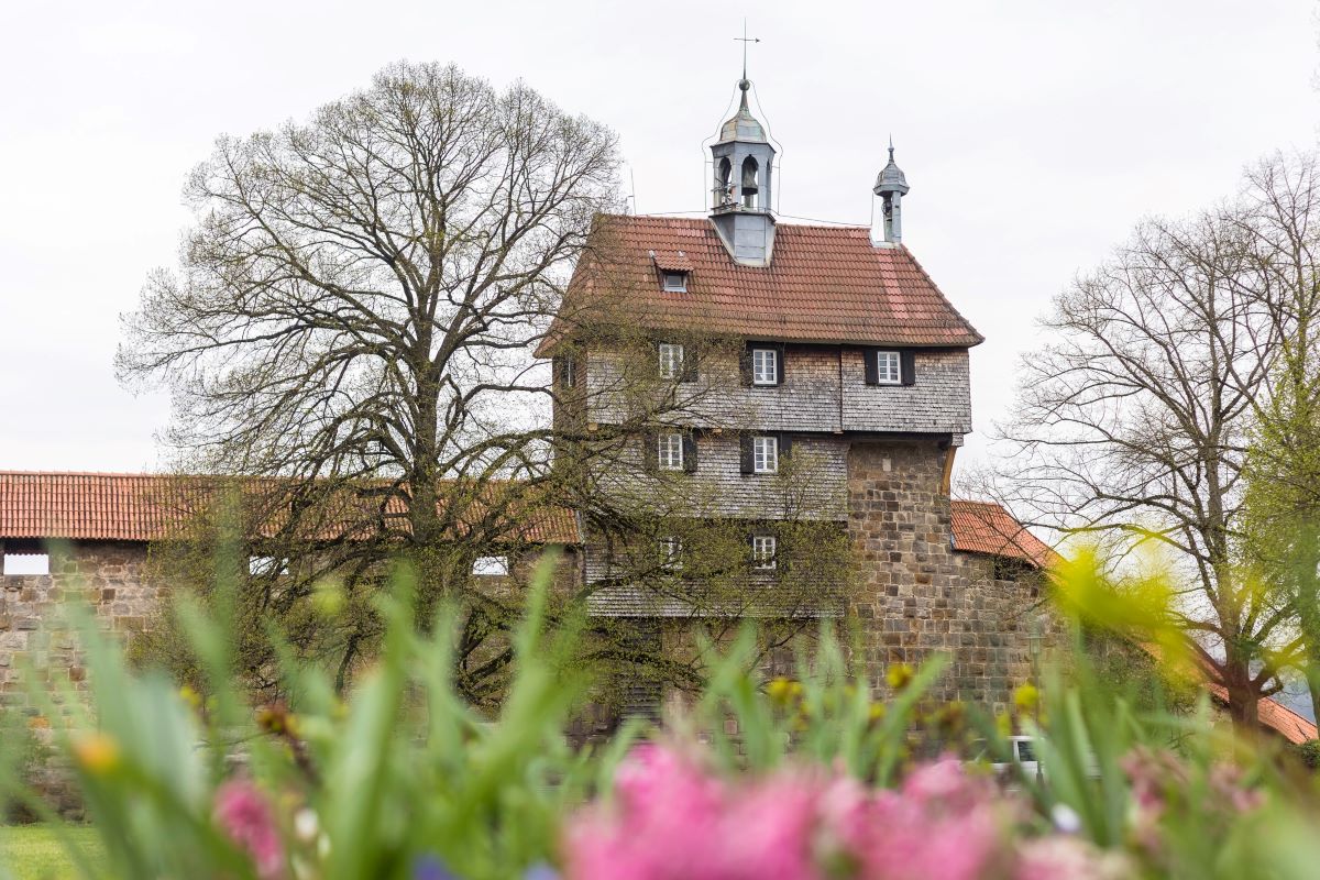Hochwacht von Norden © Thomas Wagner Schindelbedecktes Gebäude mit Glockentürmchen auf einer Mauer. Im Vordergrund Blumen.