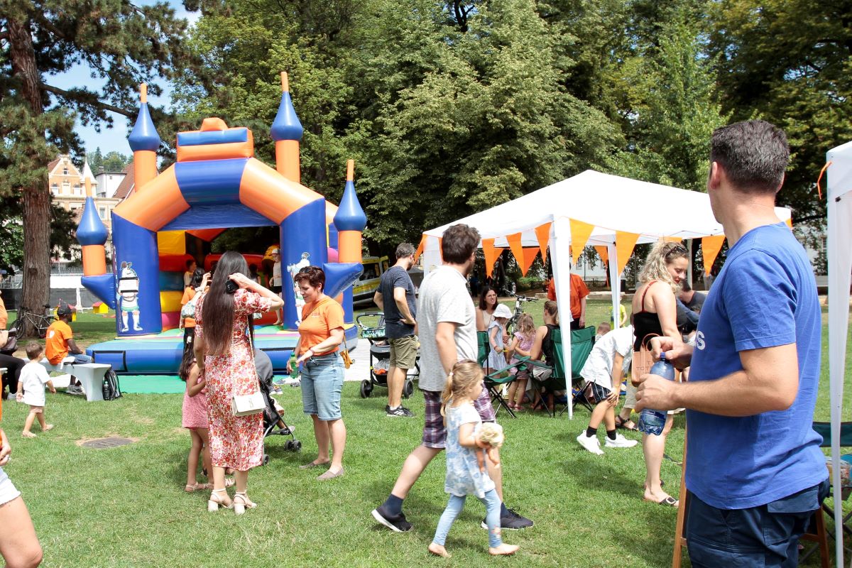 Menschen feiern bei sonnigem Wetter auf einer Wiese mit Hüpfburg und Pavillons; Kinder spielen, Erwachsene unterhalten sich.