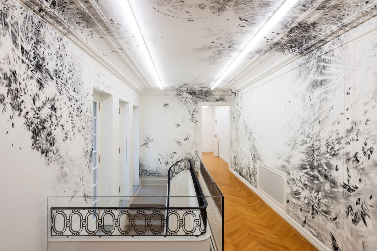 Blick in ein helles Treppenhaus mit Parkettboden und Jugendstil-Treppengeländer. Die Wände sind bis zur Stuckdecke mit schwarz-weißen Zeichnungen übersäht.