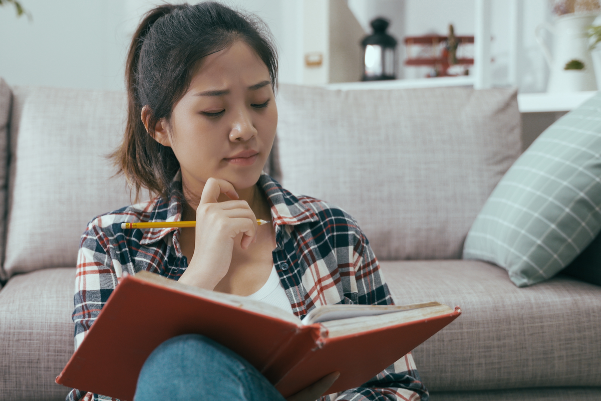 Fotografie einer jungen Asiatin, die im Wohnzimmer vor der Couch sitzt und nachdenklich in ein Buch schaut