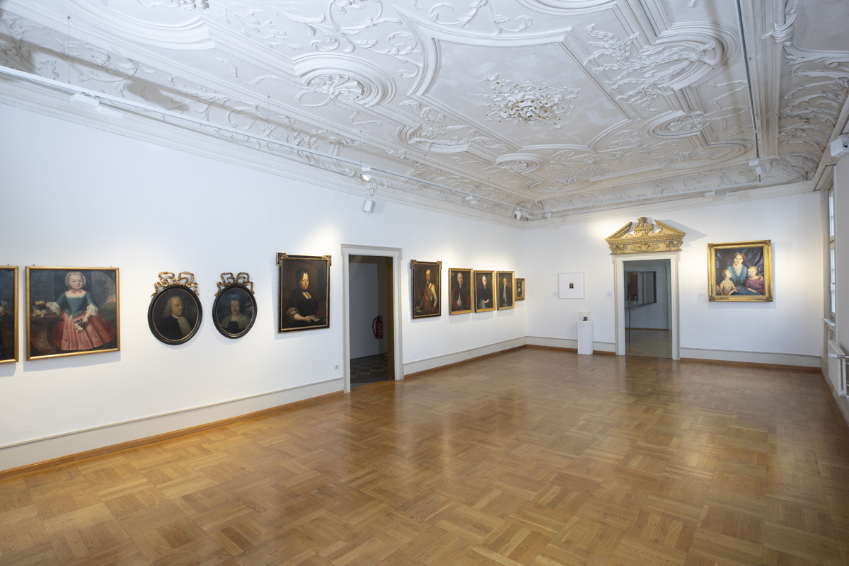 Saal mit Stuckdecke und Gemälden an den Wänden.