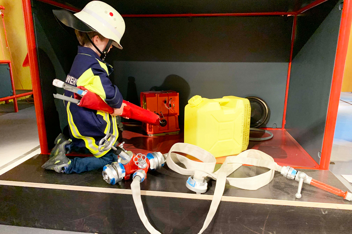 Ausstellungsraum "Wer rennt, wenn's brennt?": Kind in Feuerwehruniform spielt mit Löschschlauch. Ausstellungsraum "Wer rennt, wenn's brennt?": Kind in Feuerwehruniform spielt mit Löschschlauch.