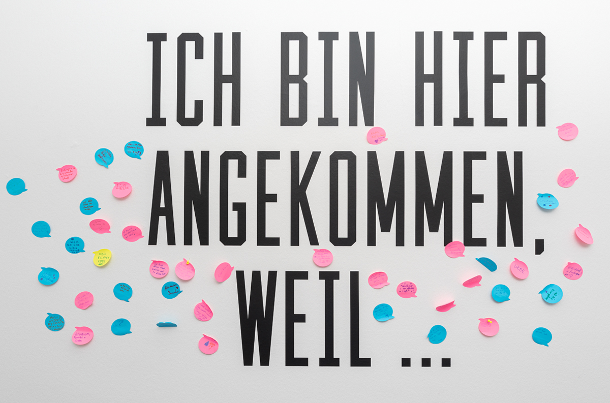 Beschriftung auf einer Wand: "ICh bin hier angekommen, weil ...". Besucherinnen und Besucher haben beschriftete Zettel danebengeklebt.