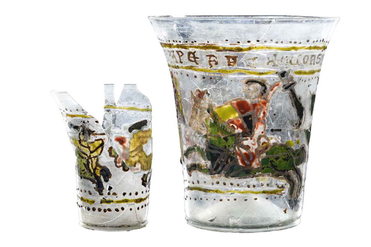 Zwei verschieden große Glasbecher, die aus Bruchstücken zusammengesetzt sind. Sie sind mit kämpfenden Reitern und Bordüren bemalt.