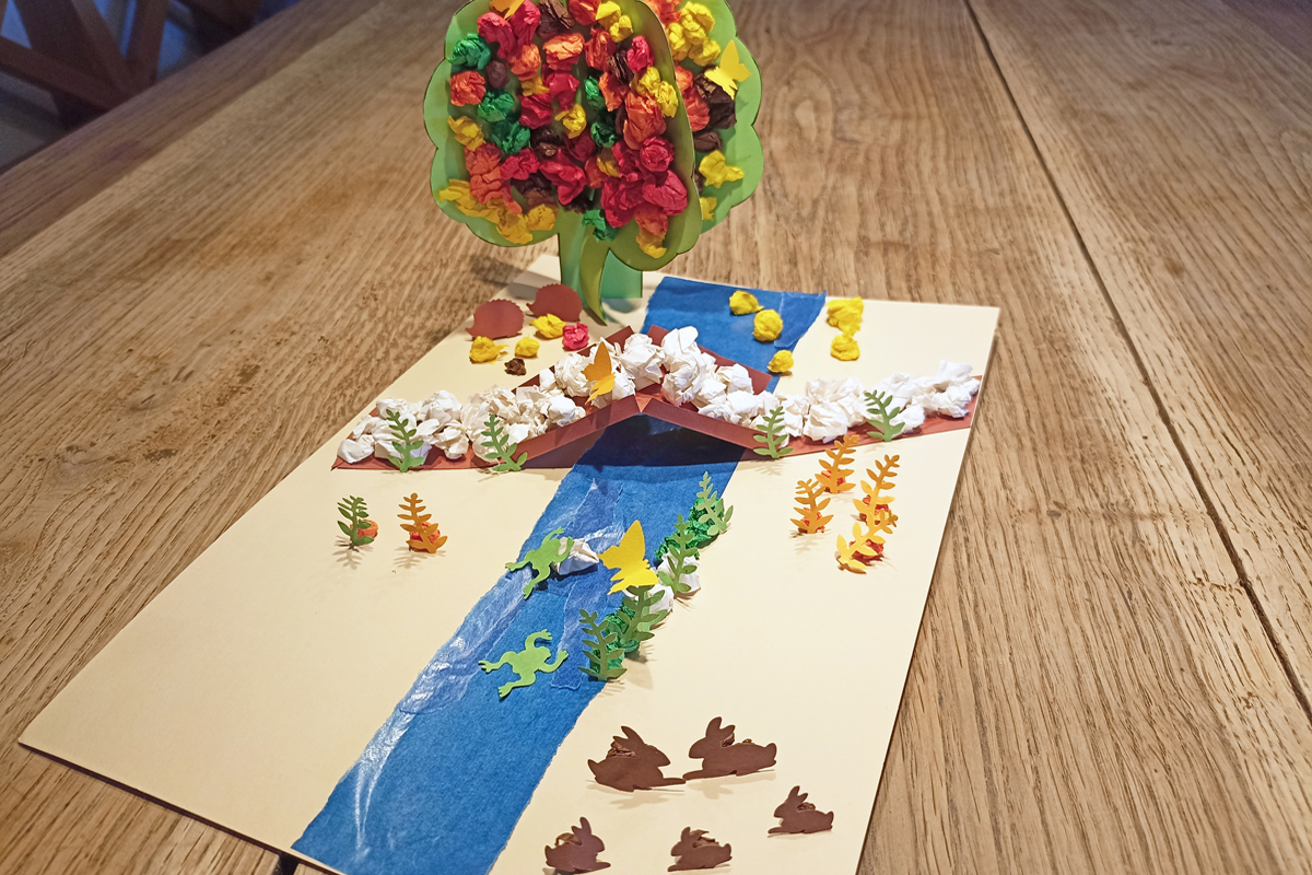 Kreative Arbeit: Aus farbigem papieren gestaltete Herbstalndschat mit einem Baum mit Früchten, einem Nach mit Brücke, Hasen und Fröschen.