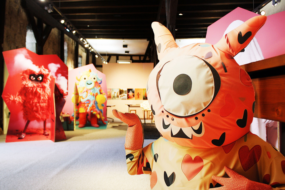 Ausstellungsraum mit großen bunten Figuren. Im Vordergrund hebt einen Stofffigur ihre Hand.
