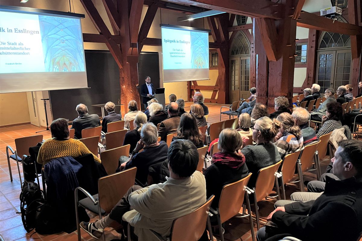 gut gefüllter Veranstaltungssaal in Reihenbestuhlung, vorne Rednerpult mit Redner, zwei Leinwände zeigen Bildschirmpräsentation